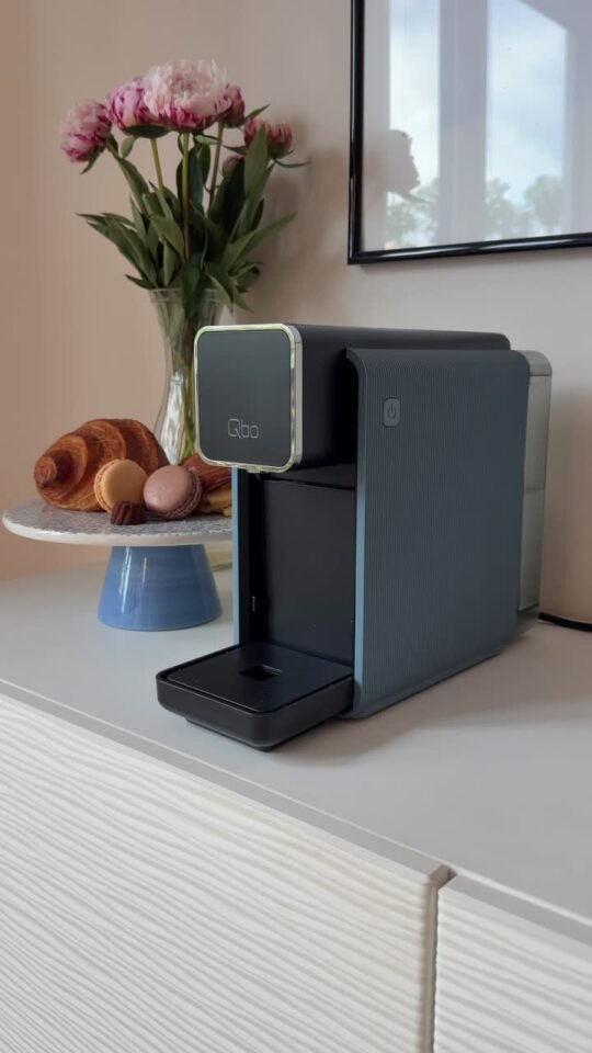 Die Tchibo-Kaffeemaschine Qbo auf einem Sideboard, daneben Pfingstrosen, Croissants und Macarons